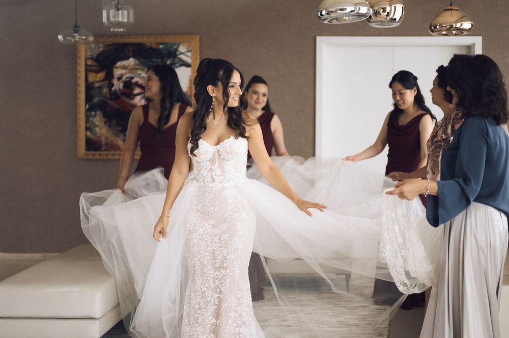 Bride in lace wedding gown surrounded by bridesmaids in burgundy dresses helping arrange her veil indoors.