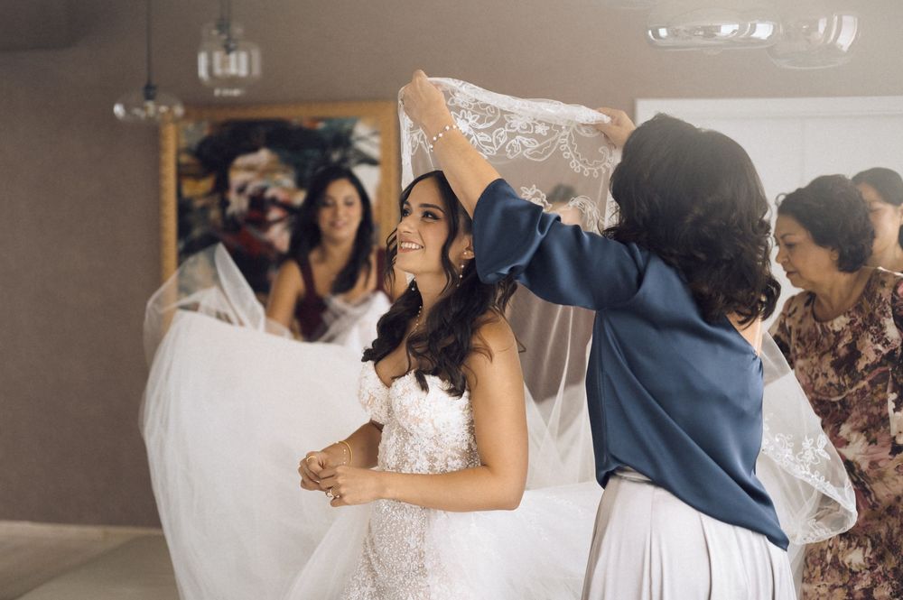 Bride smiling as bridesmaids help arrange her lace wedding veil during bridal preparations indoors.
