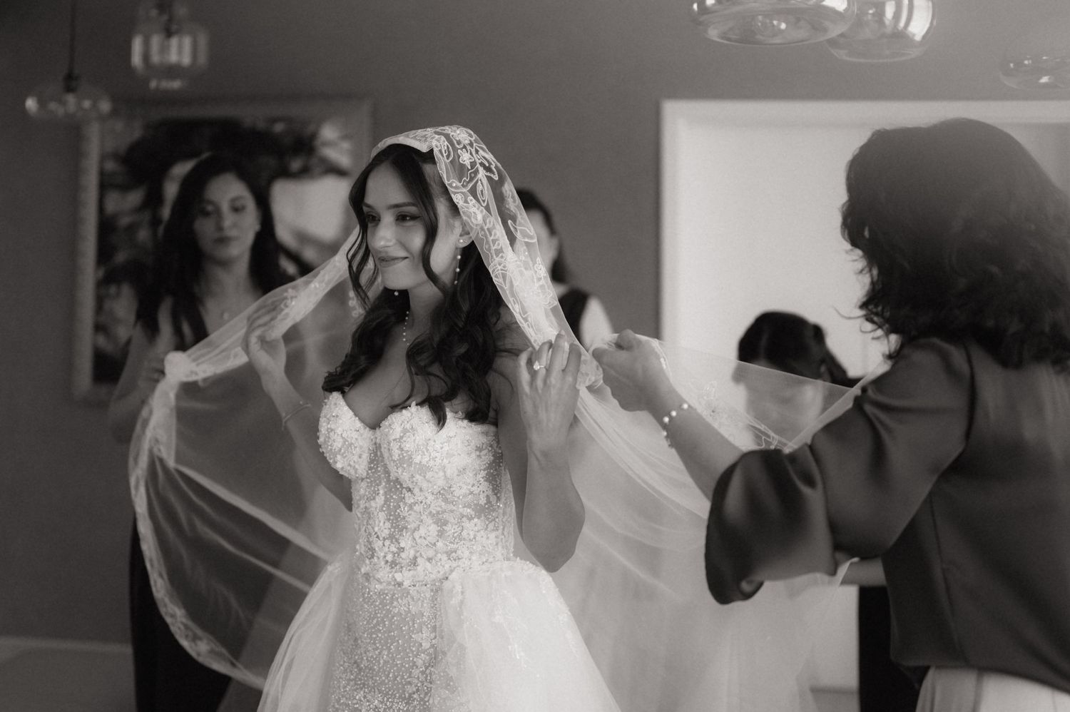 Black and white photo of a bride having her veil placed by helpers in an elegant wedding preparation scene.