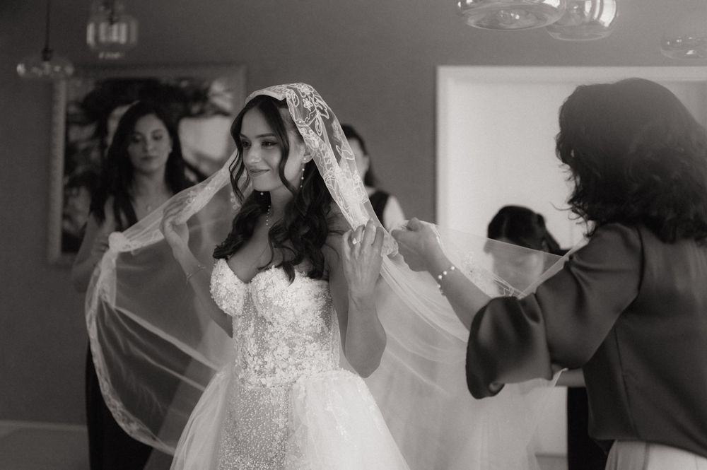 Black and white photo of a bride having her veil placed by helpers in an elegant wedding preparation scene.