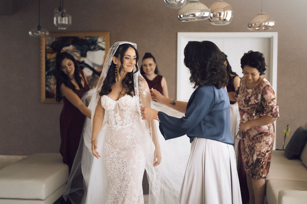 Bride in lace wedding gown with veil being assisted by bridesmaids and family in elegant bridal suite.