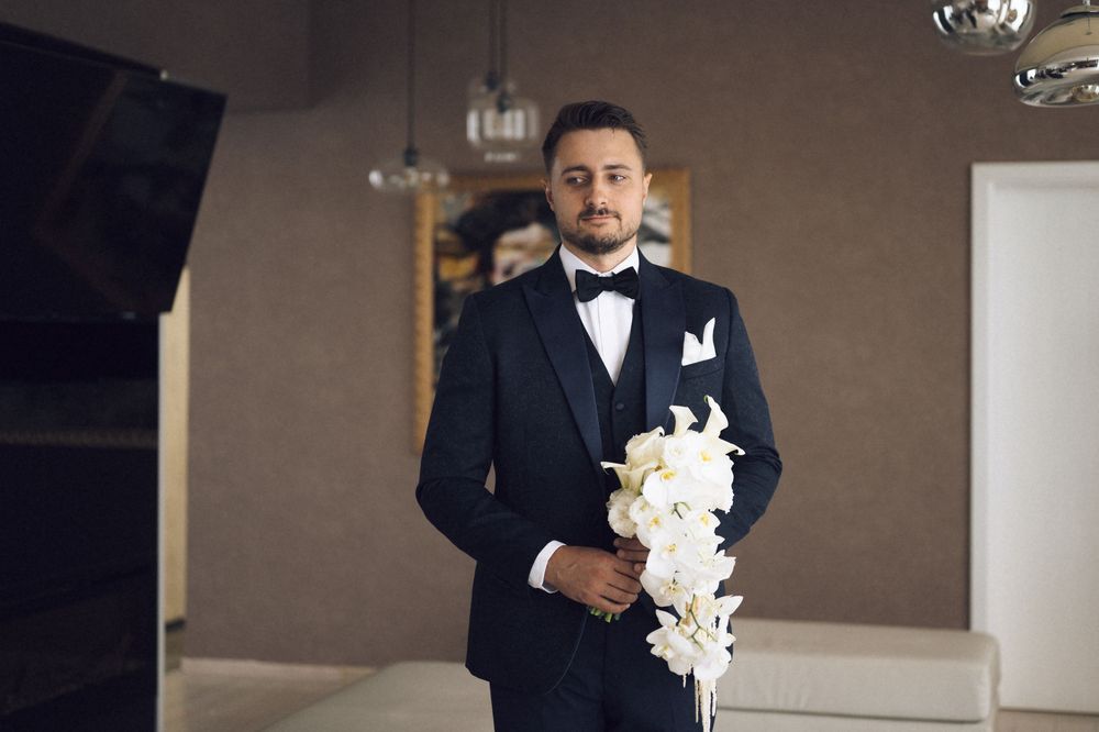 Elegant groom in black tuxedo holding white orchid bouquet standing in stylish wedding venue.