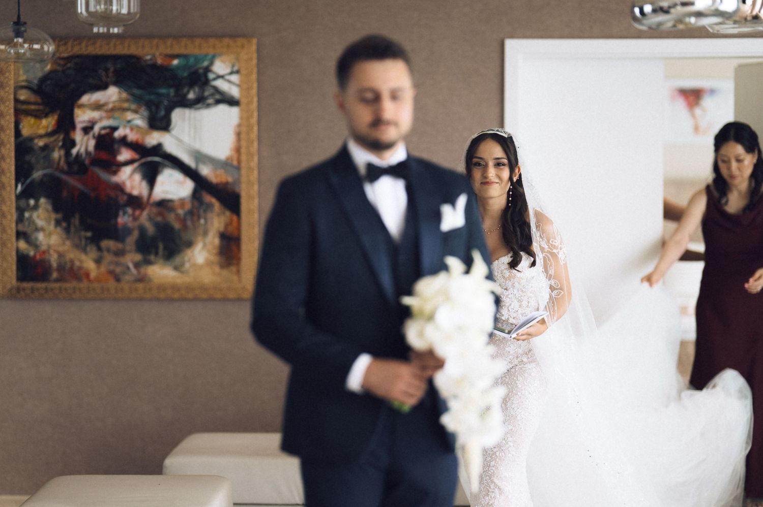 A bride in a white lace gown holds a bouquet as a groom in a tuxedo stands in the foreground at a wedding.