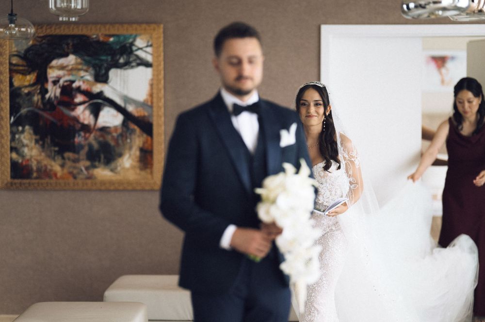 A bride in a white lace gown holds a bouquet as a groom in a tuxedo stands in the foreground at a wedding.