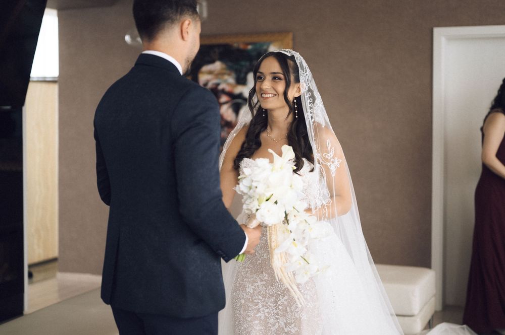 Bride in a beaded gown and veil holds white bouquet, smiling at groom in navy suit during wedding ceremony.