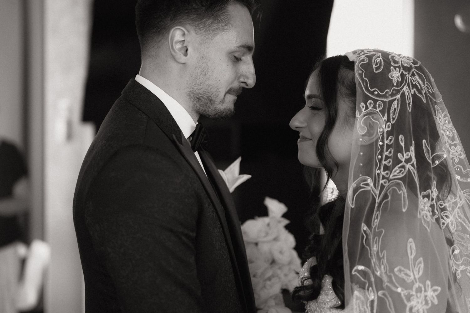 Black and white wedding photo of bride in lace veil and groom in tuxedo sharing an intimate moment.