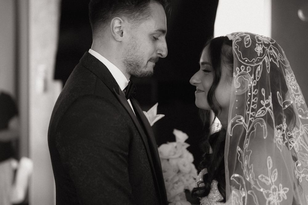 Black and white wedding photo of bride in lace veil and groom in tuxedo sharing an intimate moment.