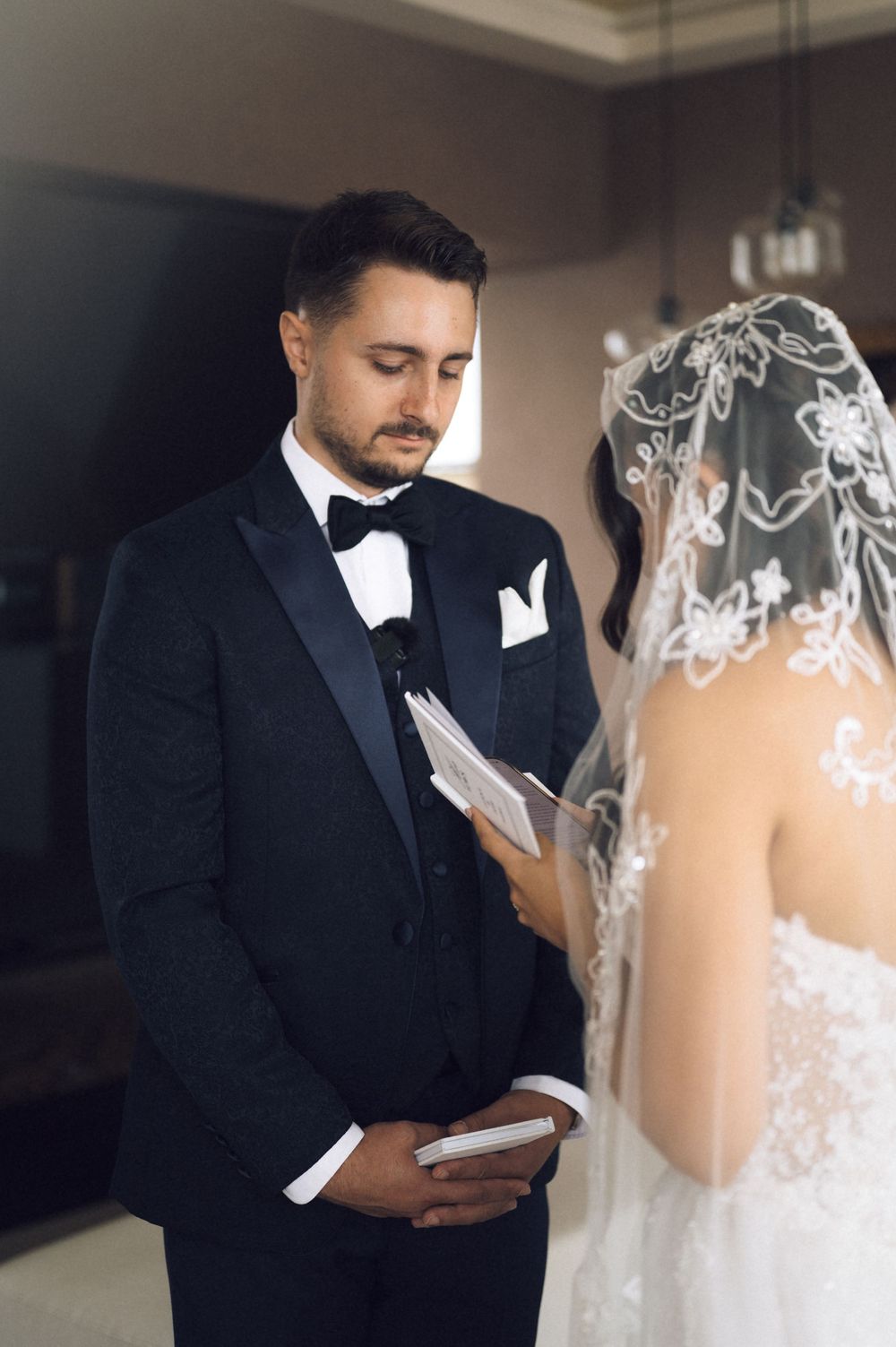 Groom in black tuxedo reads wedding vows while bride in lace dress and veil stands opposite him indoors.