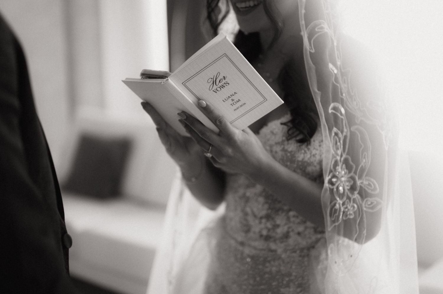 Bride in lace wedding dress and veil reading vows from a small book during wedding ceremony, black and white photo.
