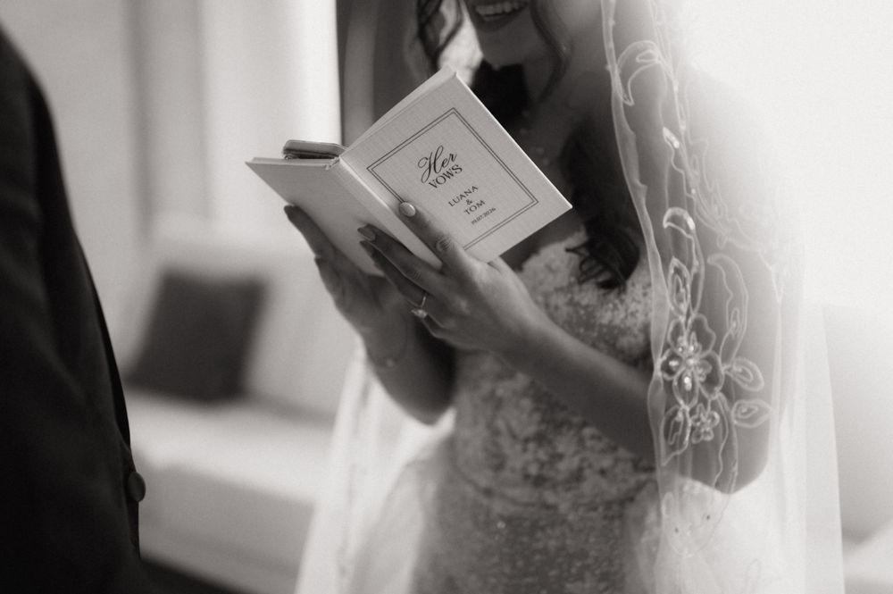 Bride in lace wedding dress and veil reading vows from a small book during wedding ceremony, black and white photo.