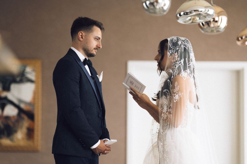 Bride in white gown and veil reads wedding vows to groom in black tuxedo during intimate ceremony.