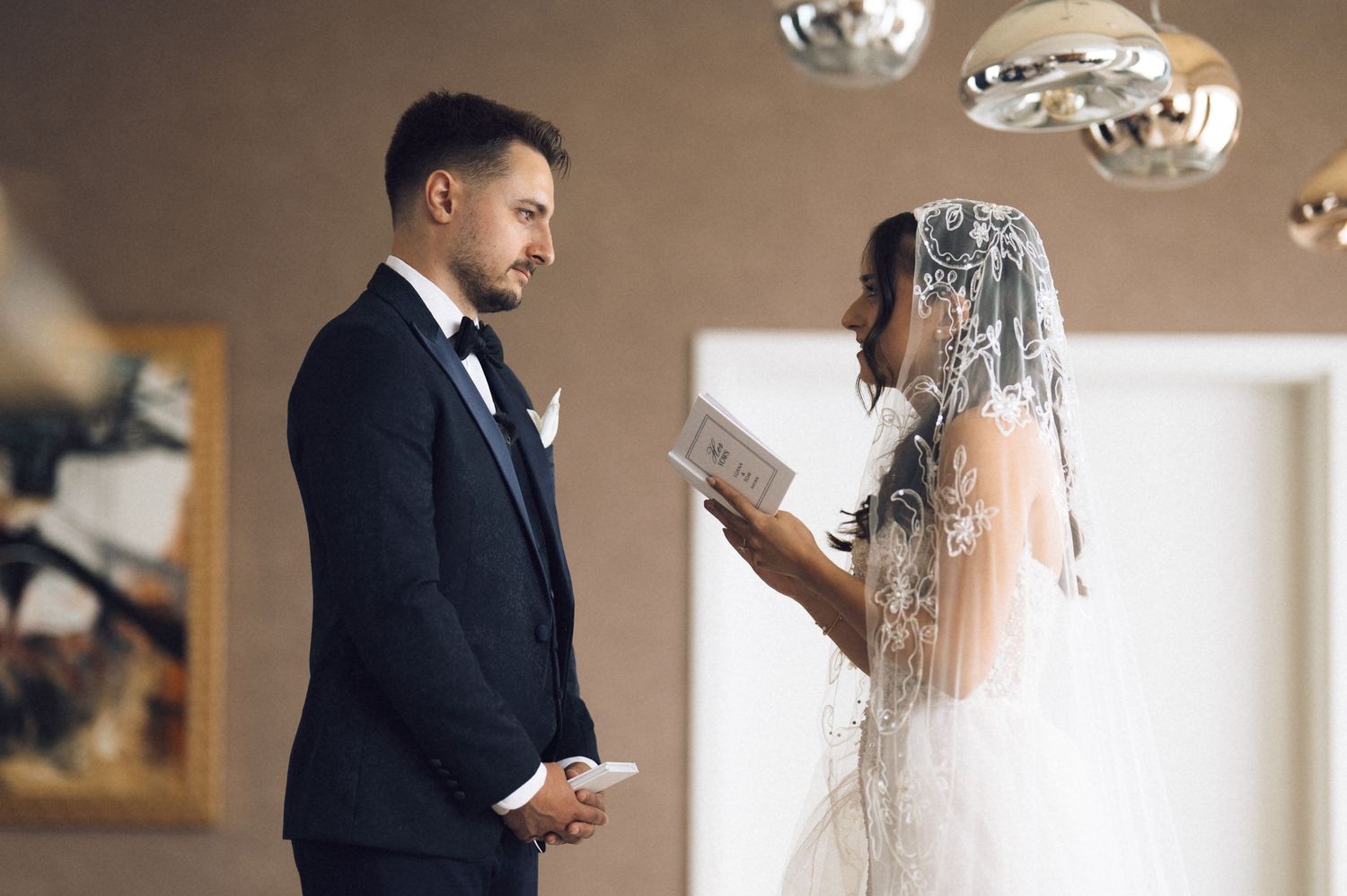 Bride in white gown and veil reads wedding vows to groom in black tuxedo during intimate ceremony.