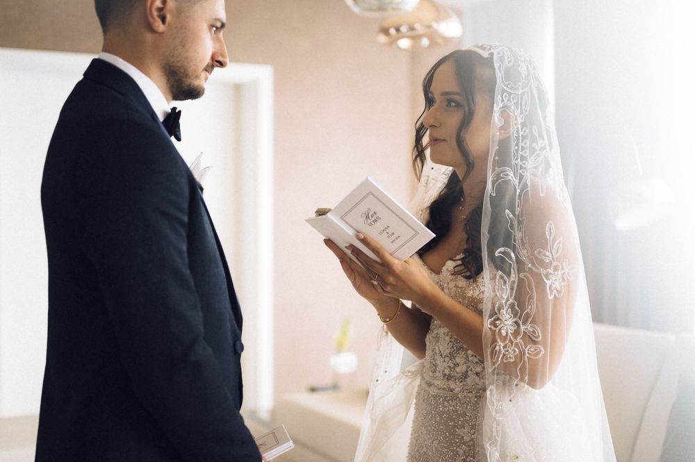 Bride in lace wedding dress and veil reads vows from a book while groom in black tuxedo listens during ceremony.