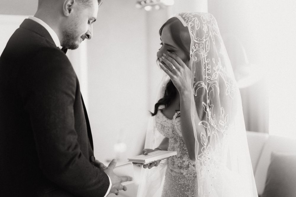 Black and white photo of emotional bride covering mouth while groom reads wedding vows during intimate ceremony.