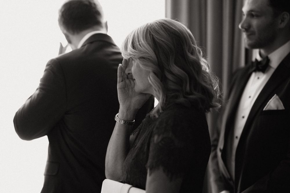 Black and white wedding photo of emotional mother covering her mouth with hand during ceremony.