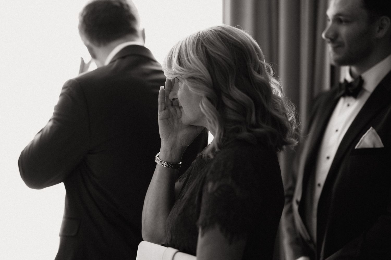 Black and white wedding photo of emotional mother covering her mouth with hand during ceremony.