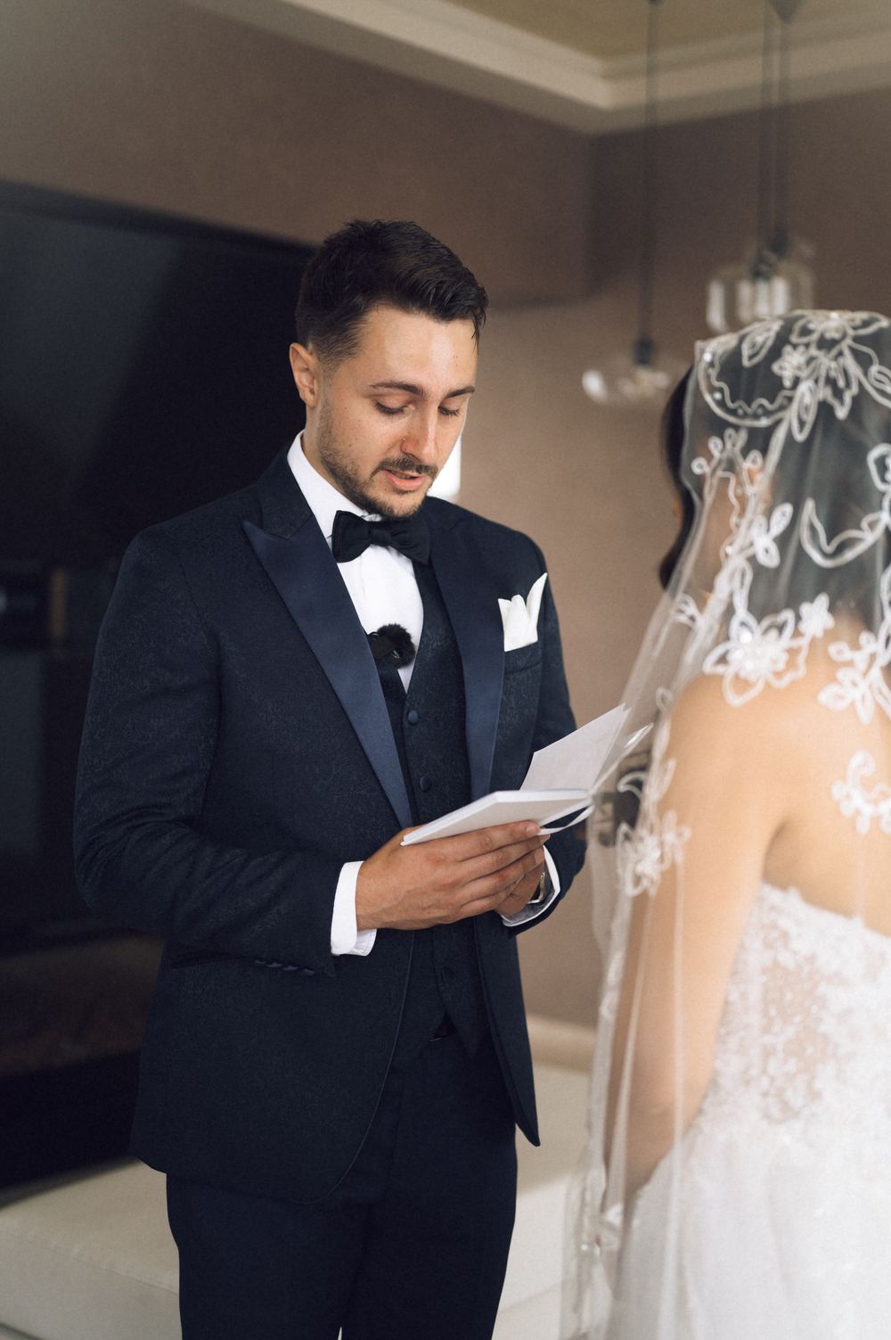 Groom in black tuxedo reads wedding vows to bride in lace dress and veil during intimate ceremony.