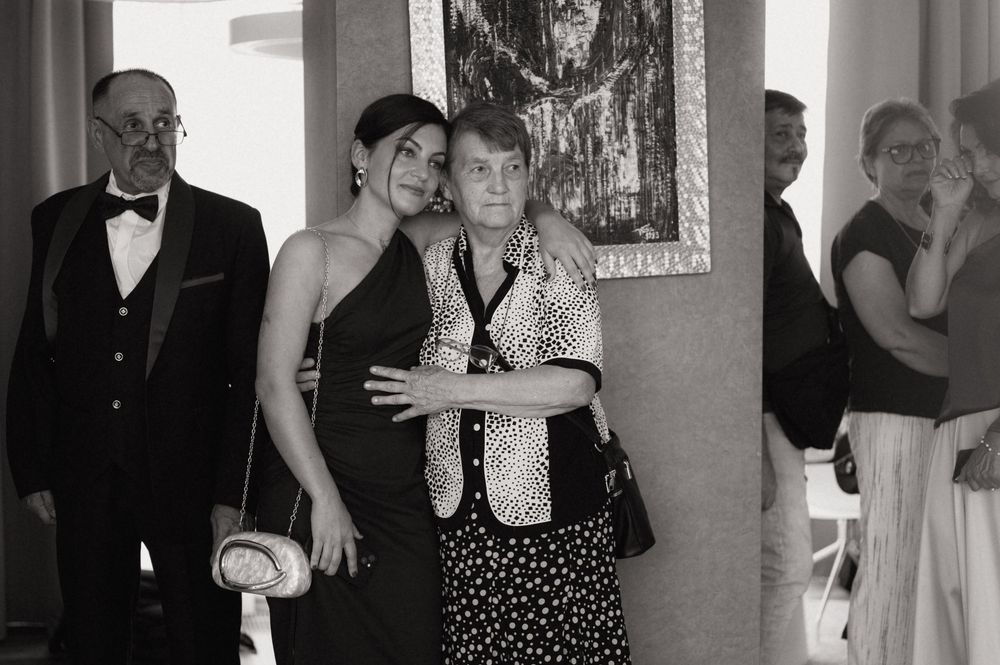 Black and white photo of two women embracing at a formal event, with an elderly man and others visible in background.
