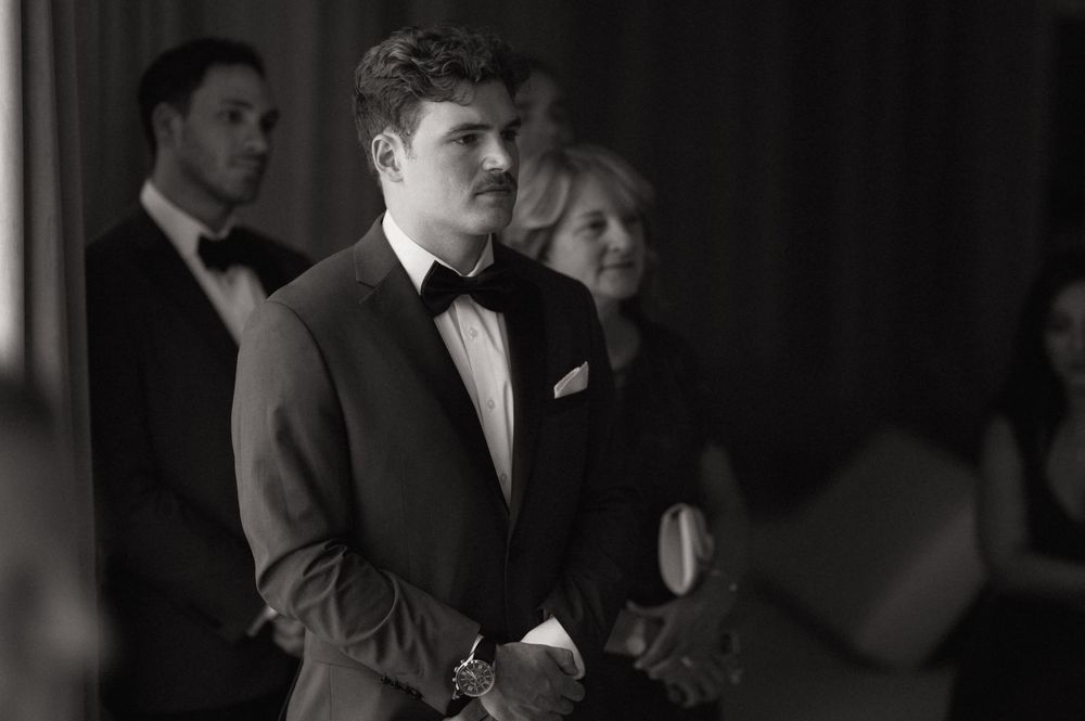 Groom in black bow tie and suit watches bride approach at wedding ceremony, black and white photo.