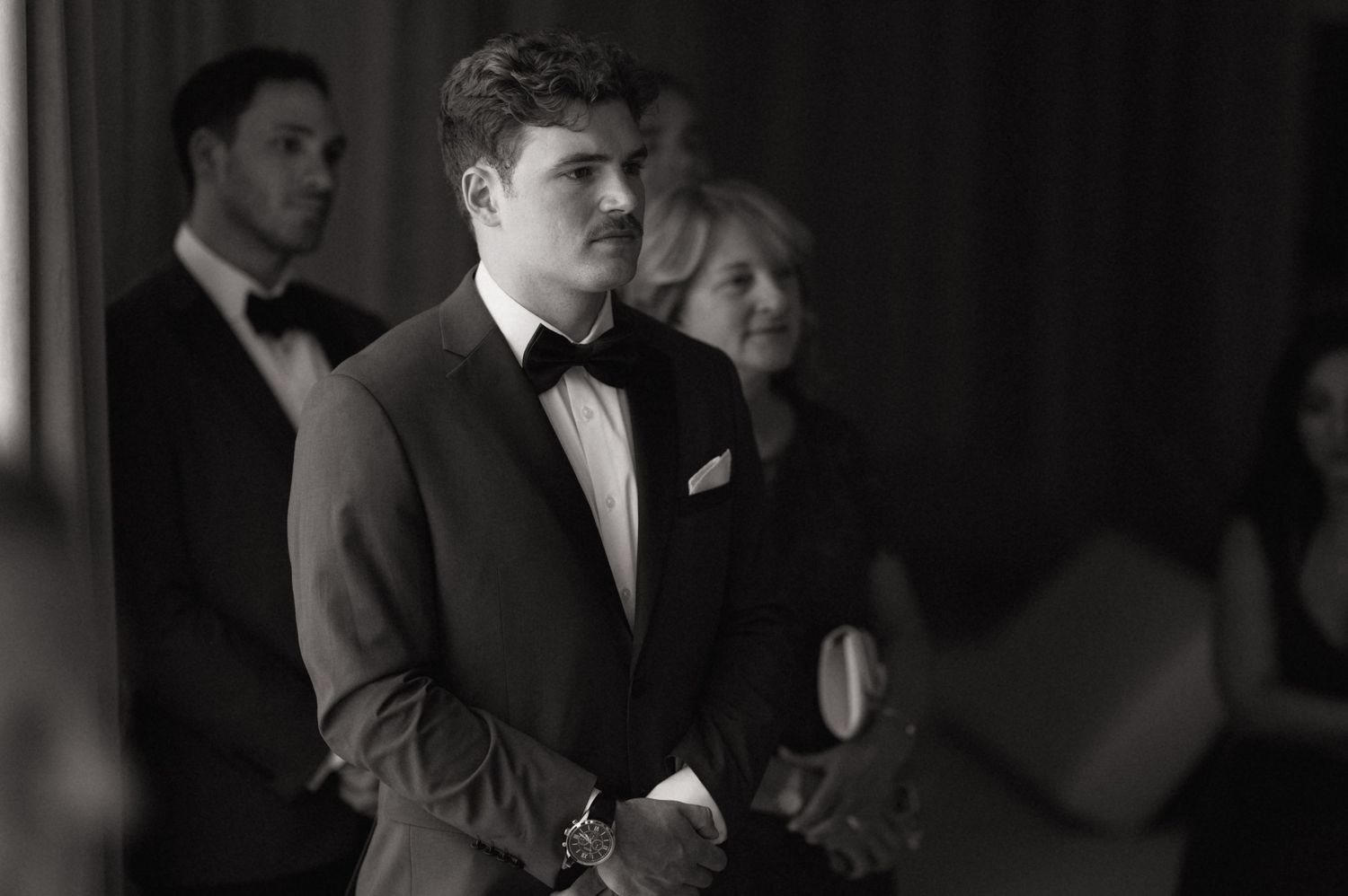 Groom in black bow tie and suit watches bride approach at wedding ceremony, black and white photo.
