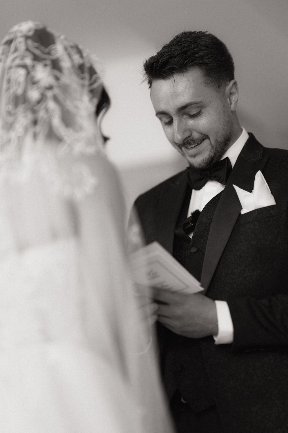 Groom in tuxedo smiling while reading wedding vows to bride in lace dress, black and white ceremony photo.