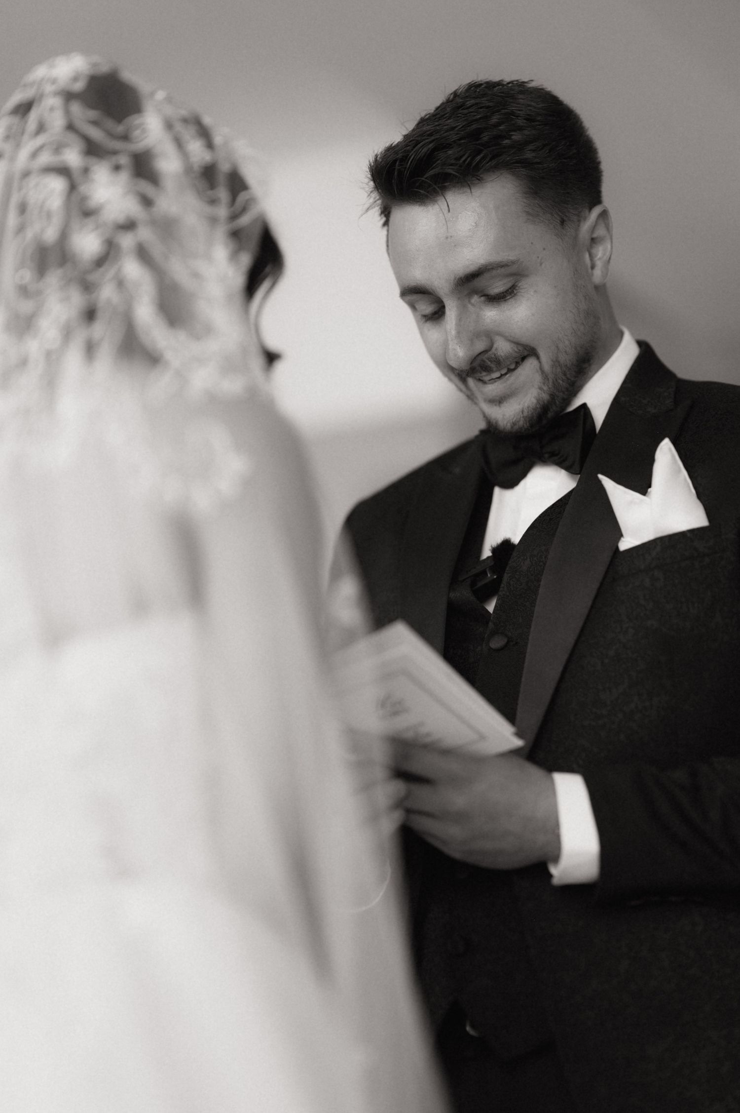 Groom in tuxedo smiling while reading wedding vows to bride in lace dress, black and white ceremony photo.