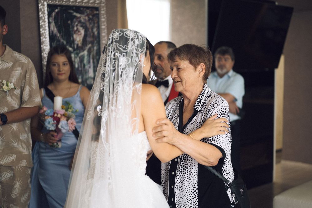 Older woman warmly embracing a bride in a white wedding dress and veil during a wedding celebration indoors.