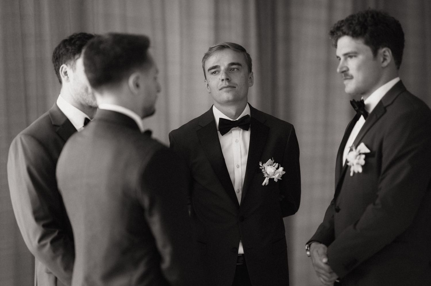 Groomsmen in black tuxedos share a candid moment before wedding ceremony in black and white photo.