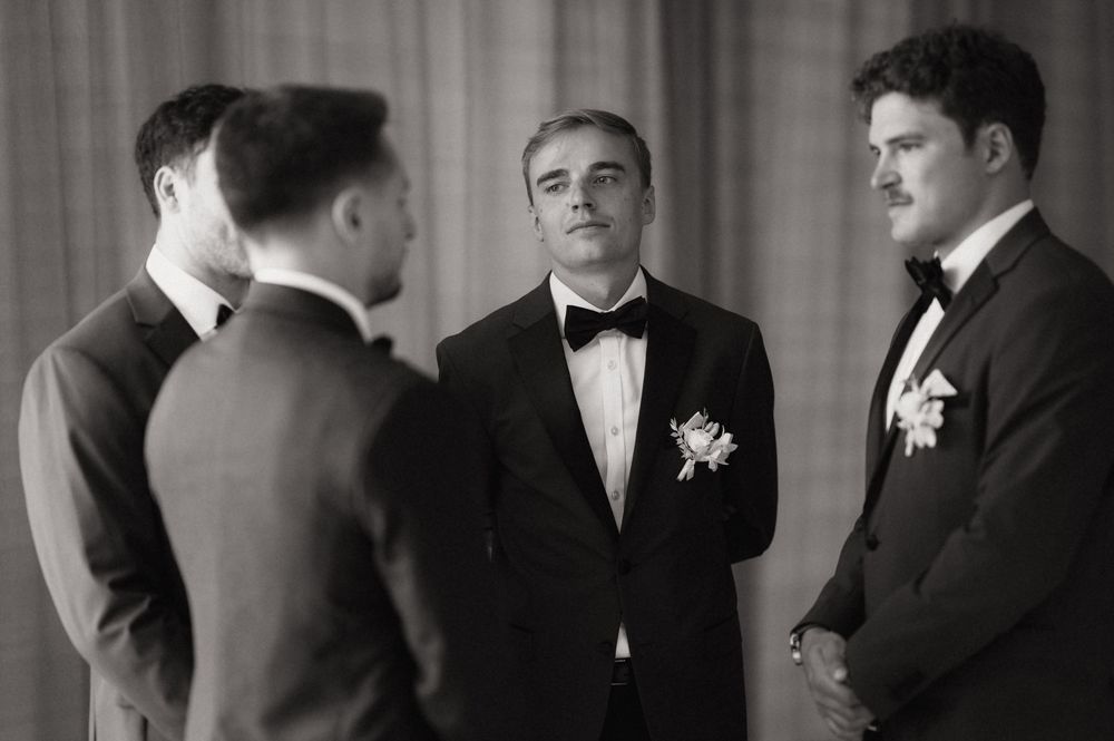 Groomsmen in black tuxedos share a candid moment before wedding ceremony in black and white photo.
