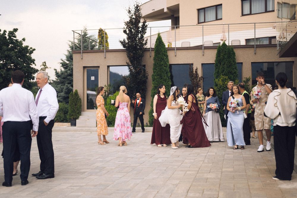 Wedding party guests in formal attire gathered outside a modern building with tall cypress trees.