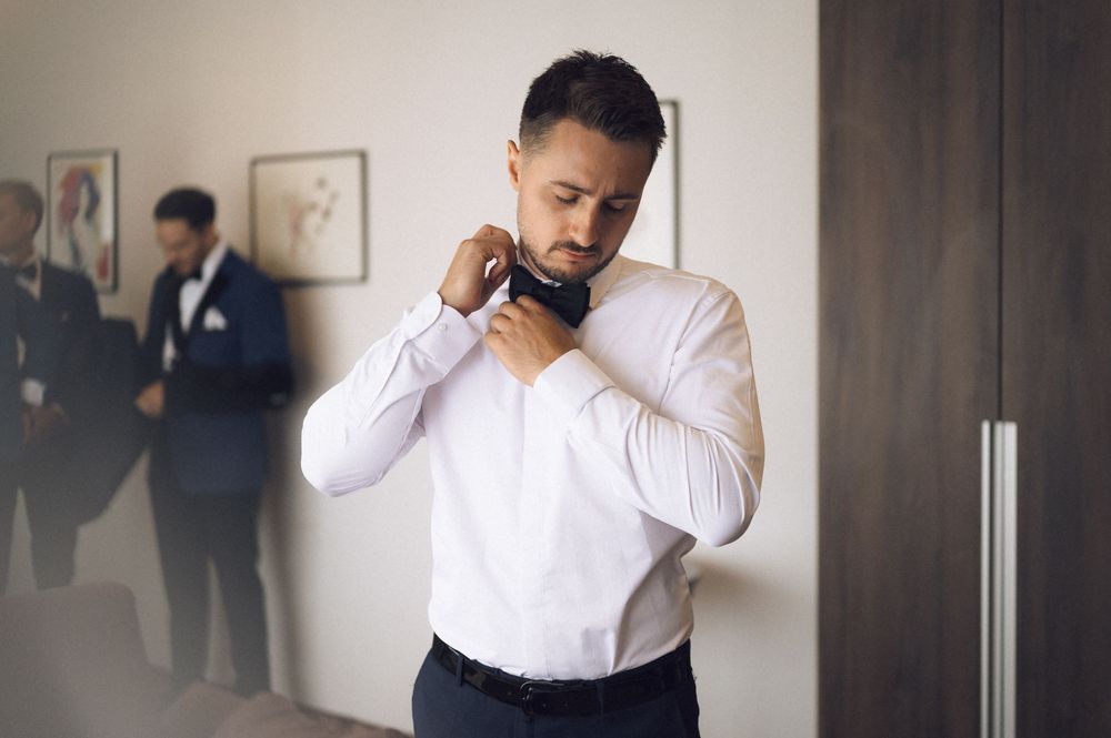 Groom in white dress shirt adjusting his navy bow tie while getting ready with groomsmen in background.