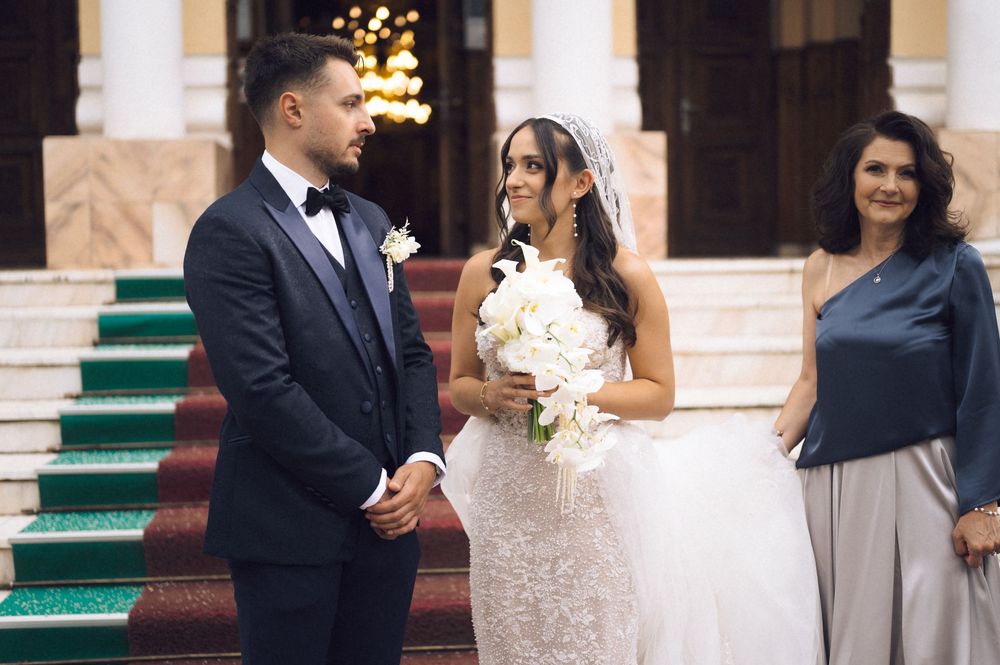 Bride and groom stand together at ornate staircase, bride holding white bouquet in lace wedding gown.