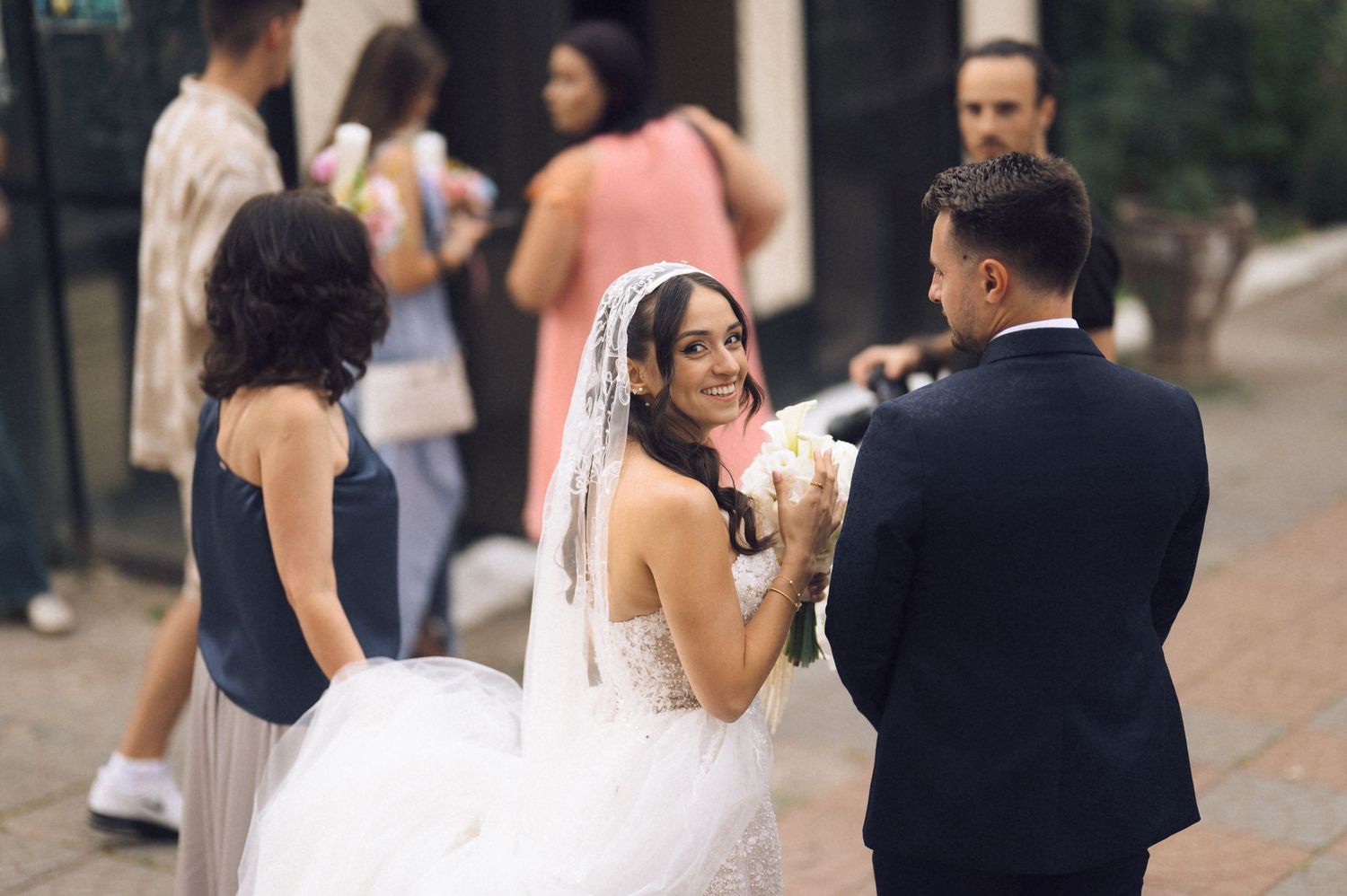 Smiling bride in white gown and veil holds bouquet while chatting with groom in black suit at outdoor wedding.
