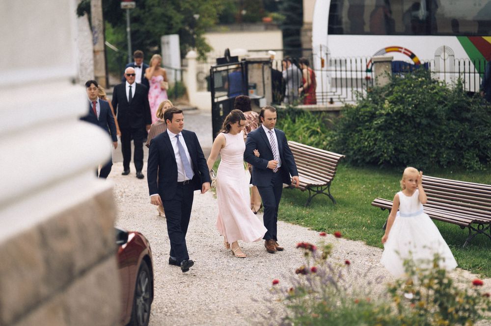 Wedding party walking outdoors, bride in white gown with guests in formal attire, flower girl leads procession.