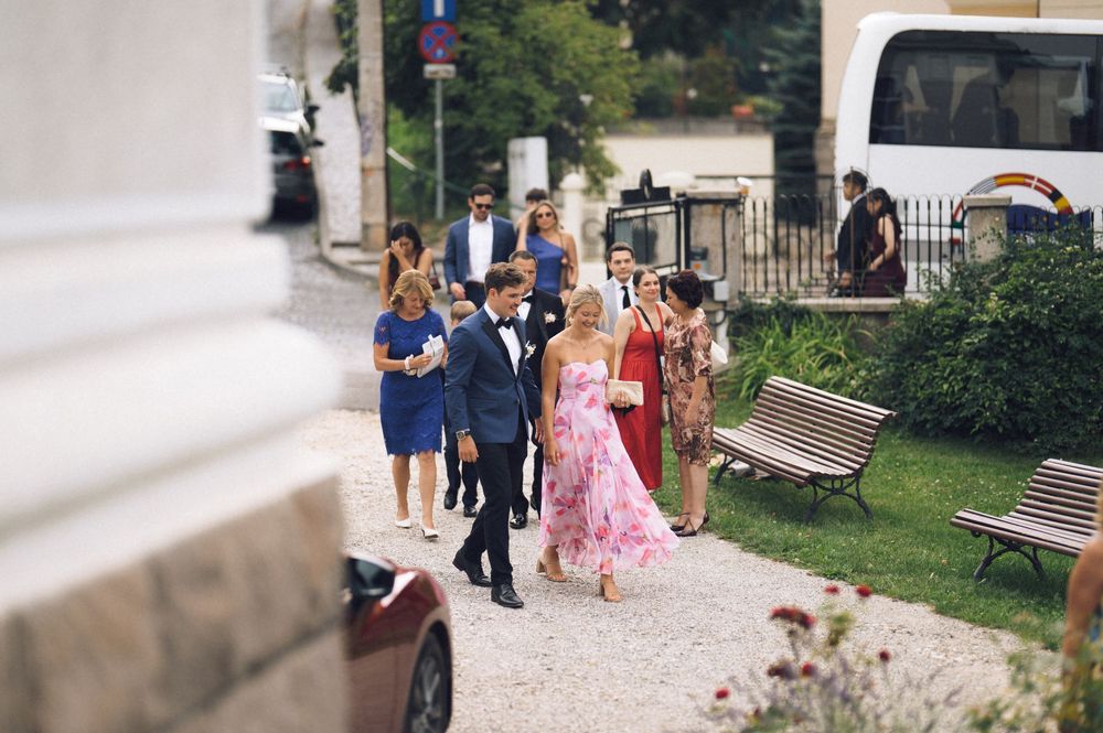Elegantly dressed wedding guests walk outdoors, featuring a woman in a pink floral dress and a man in a tuxedo.