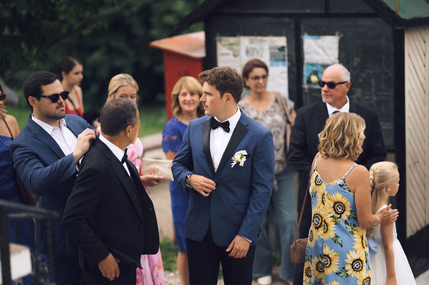 Groom in blue suit with bow tie greeting wedding guests outdoors at a summer ceremony.