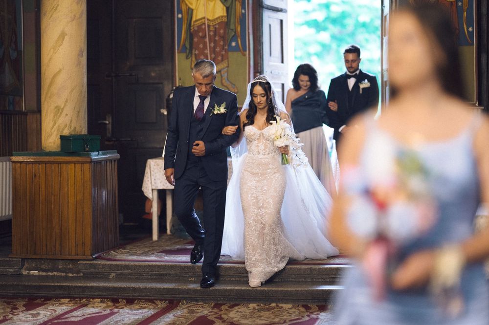 Bride walks down church aisle with father in elegant lace wedding gown, guests following behind.