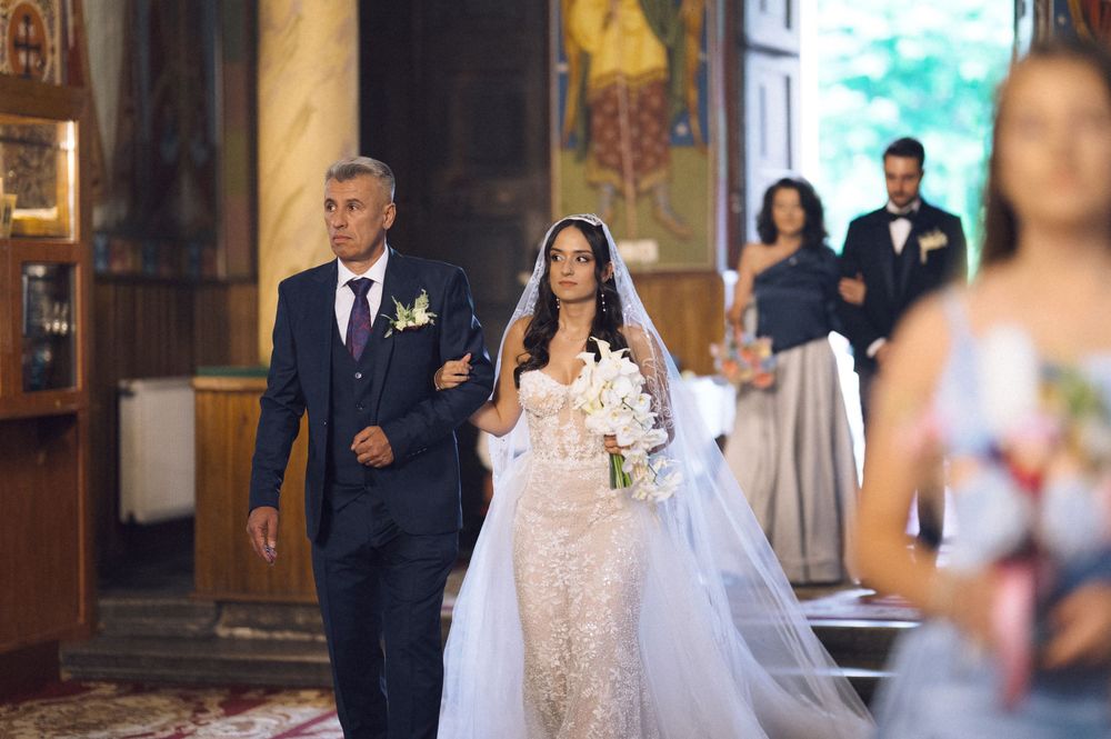 Bride in lace wedding gown walks down church aisle with father, groomsman and bridesmaid visible behind.