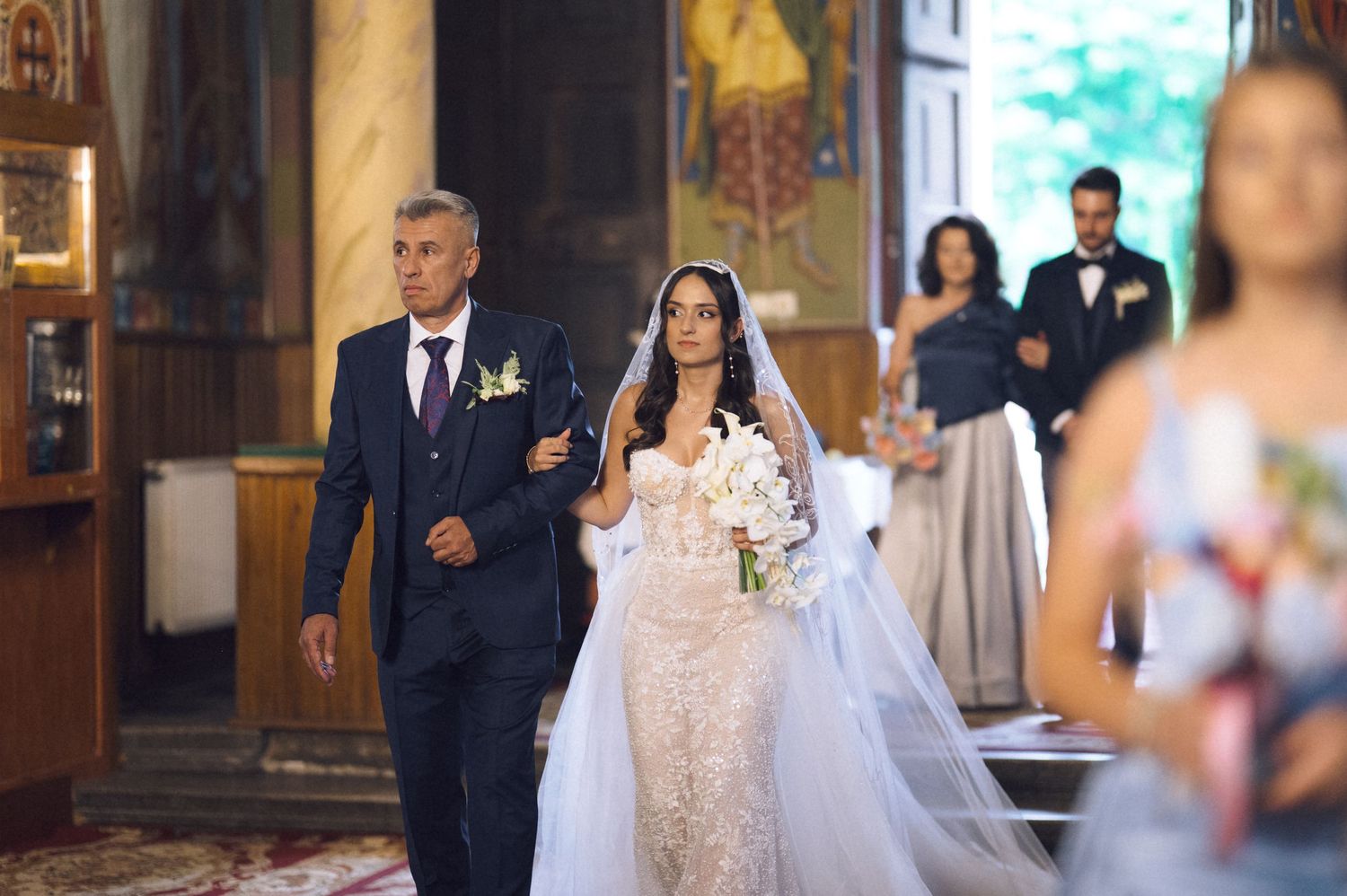 Bride in lace wedding gown walks down church aisle with father, groomsman and bridesmaid visible behind.