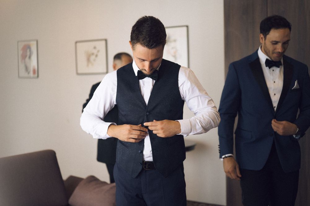 Two groomsmen in navy suits with bow ties prepare for a wedding ceremony while adjusting their formal attire indoors.