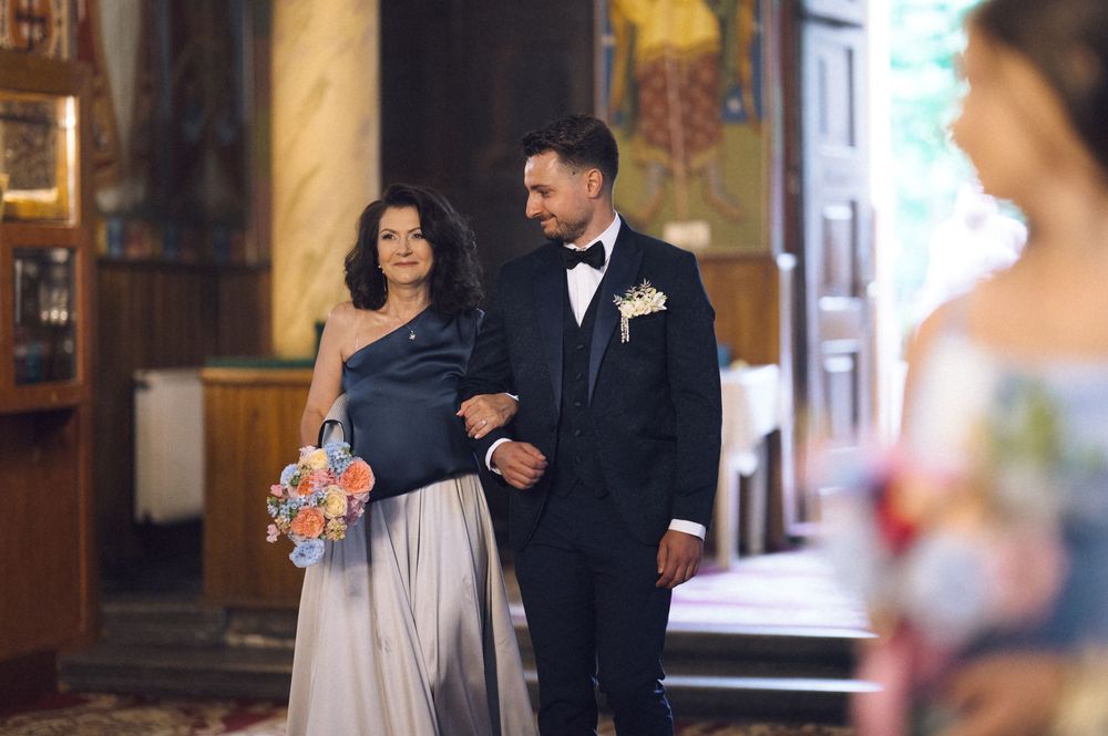 Groom in tuxedo escorts woman in blue gown down church aisle at elegant wedding ceremony.