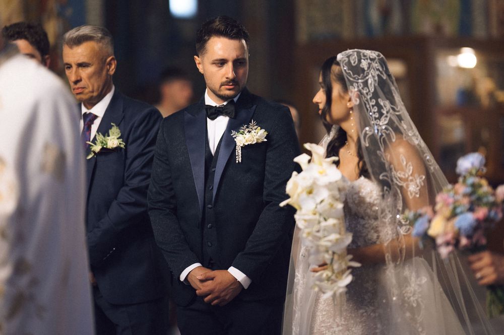 Groom in black tuxedo stands at church altar with bride in white lace gown and veil during wedding ceremony.