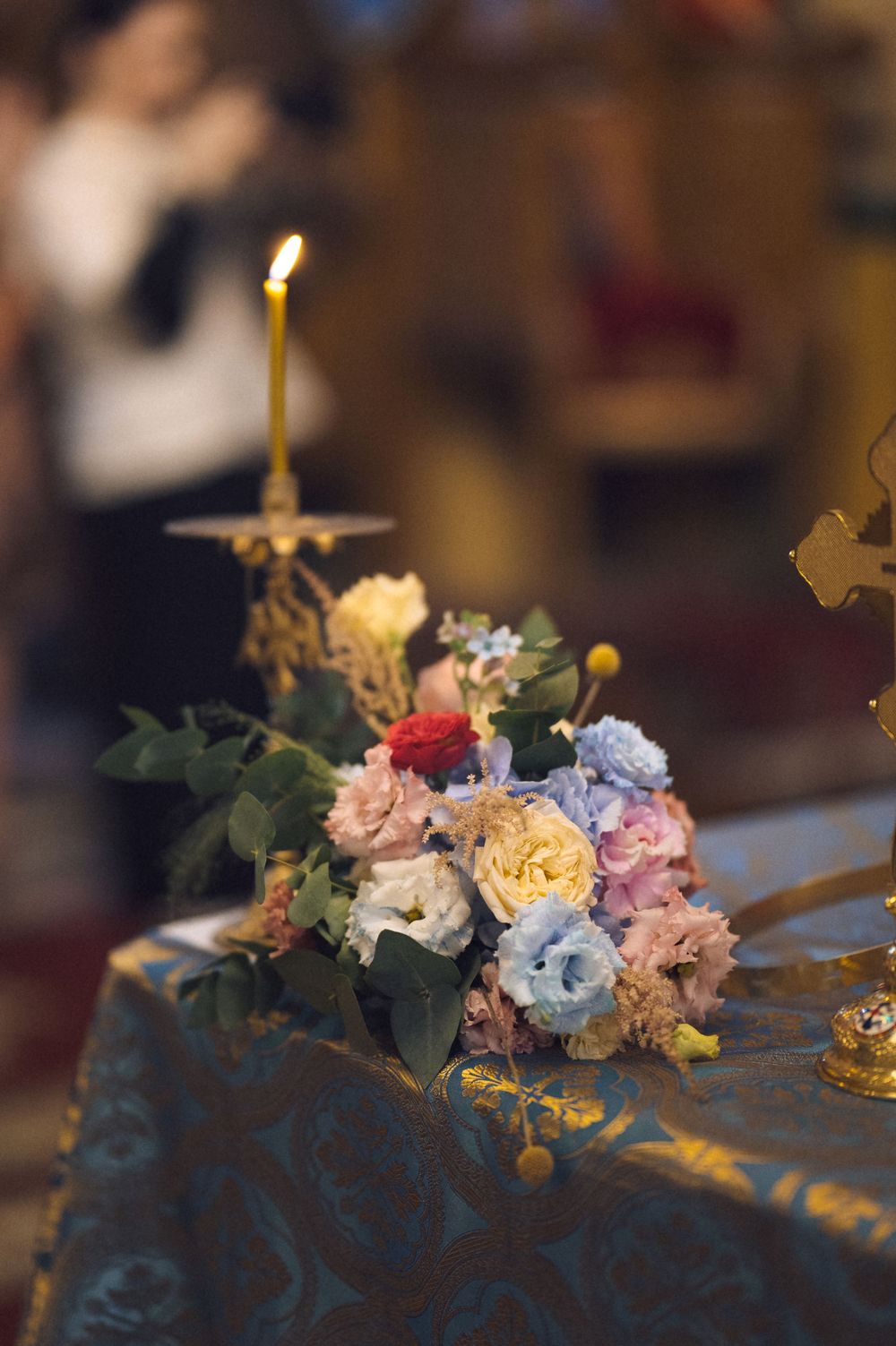 Colorful floral bouquet with roses on ornate blue fabric altar with lit candle in church ceremony setting.