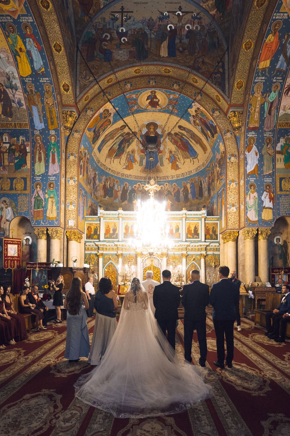 Bride and groom stand at ornate Orthodox church altar surrounded by wedding party, with stunning Byzantine frescoes and golden iconostasis.