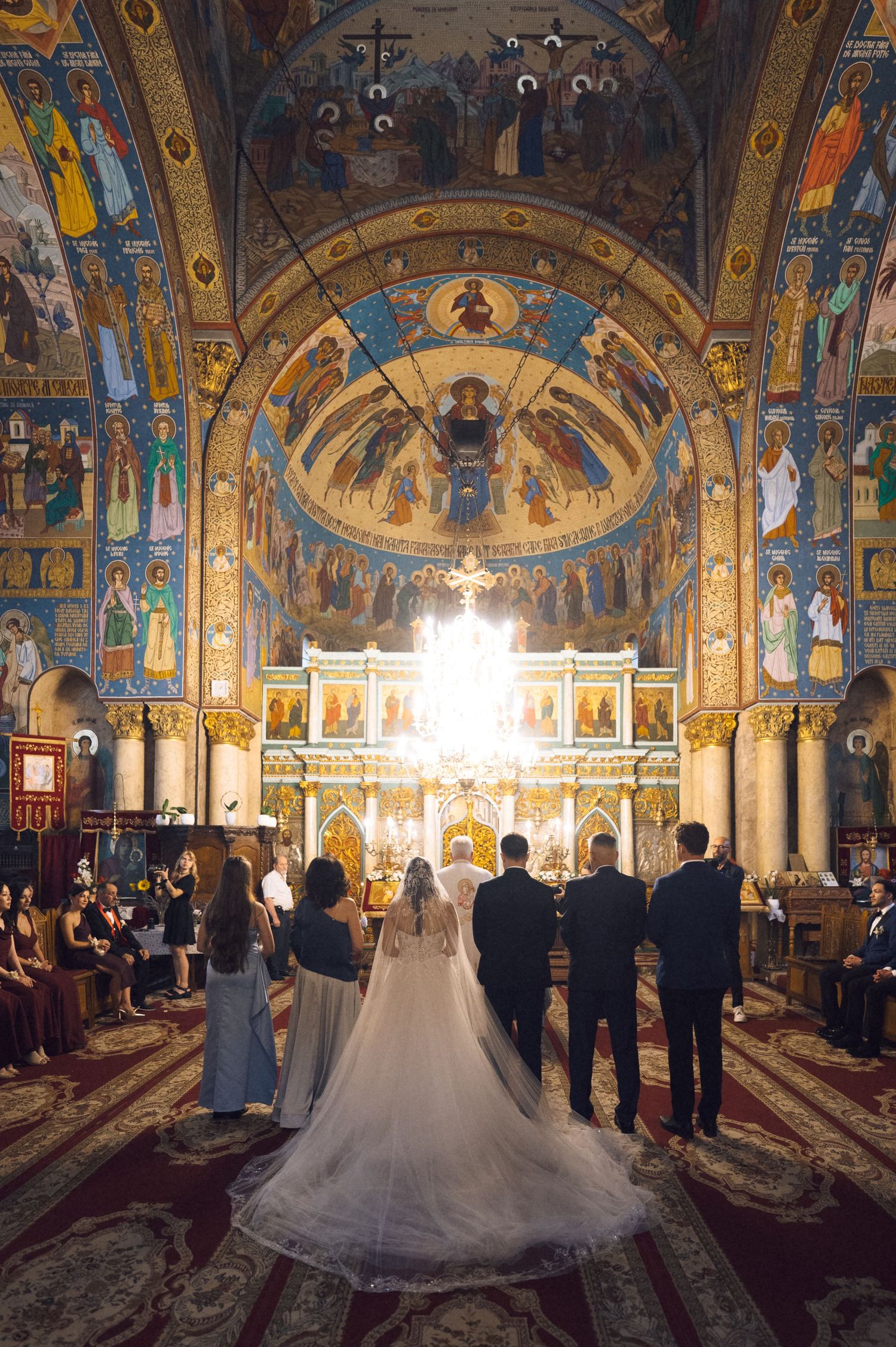 Bride and groom stand at ornate Orthodox church altar surrounded by wedding party, with stunning Byzantine frescoes and golden iconostasis.
