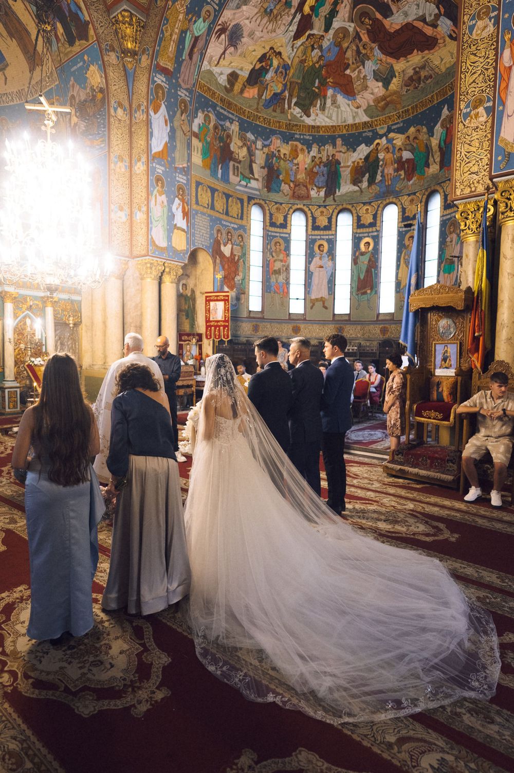 Bride in white gown with long veil at Orthodox church wedding ceremony with ornate golden mosaic interior.