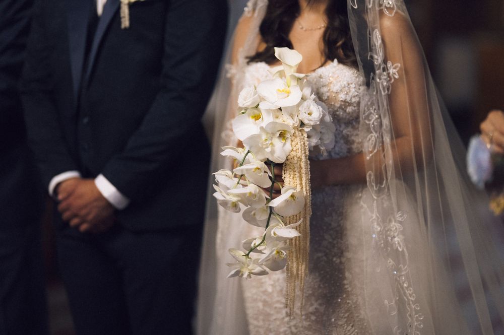 Bride in lace gown holding white orchid bouquet stands beside groom in black suit during wedding ceremony.