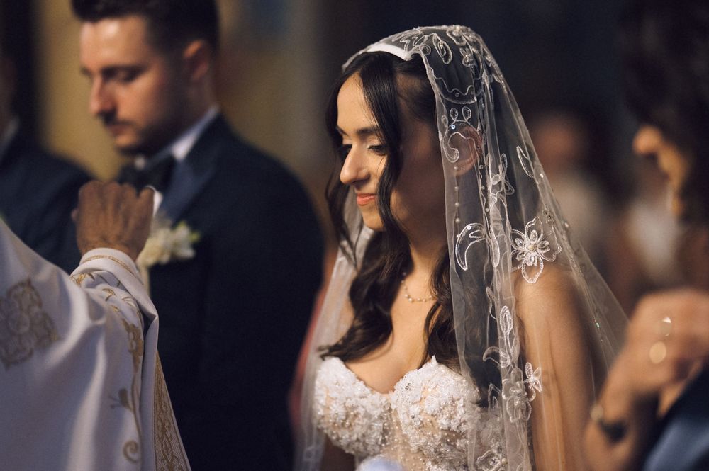 Smiling bride in lace veil and gown at church wedding ceremony with groom in background.