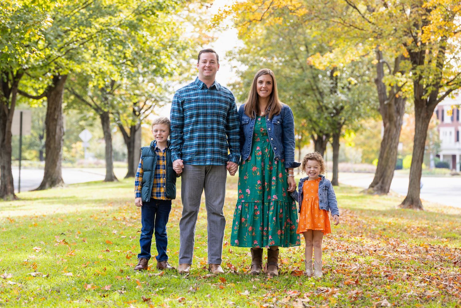 Family of four smiling outdoors in autumn park with colorful fall foliage and green grass.