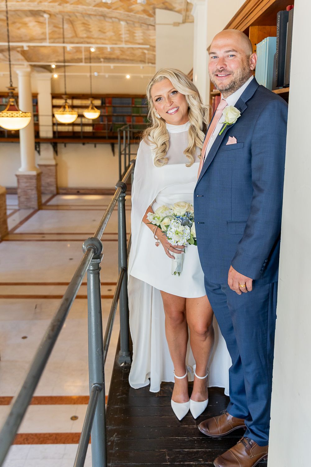Bride in white mini dress holding bouquet stands with groom in navy suit on elegant venue balcony.